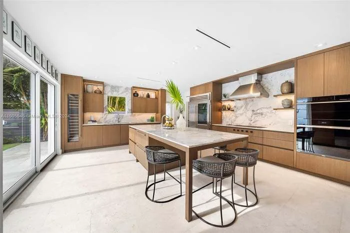 The Miami kitchen after DALOP Design: warm wood cabinetry, a marble backsplash and an island with stools